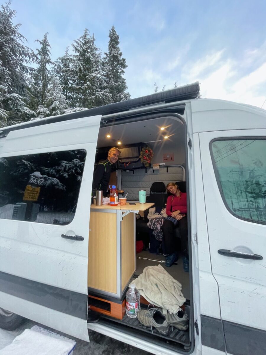 Converted Sprinter with side door open showing galley and two smiling campers, blue skies and snowy trees in the background